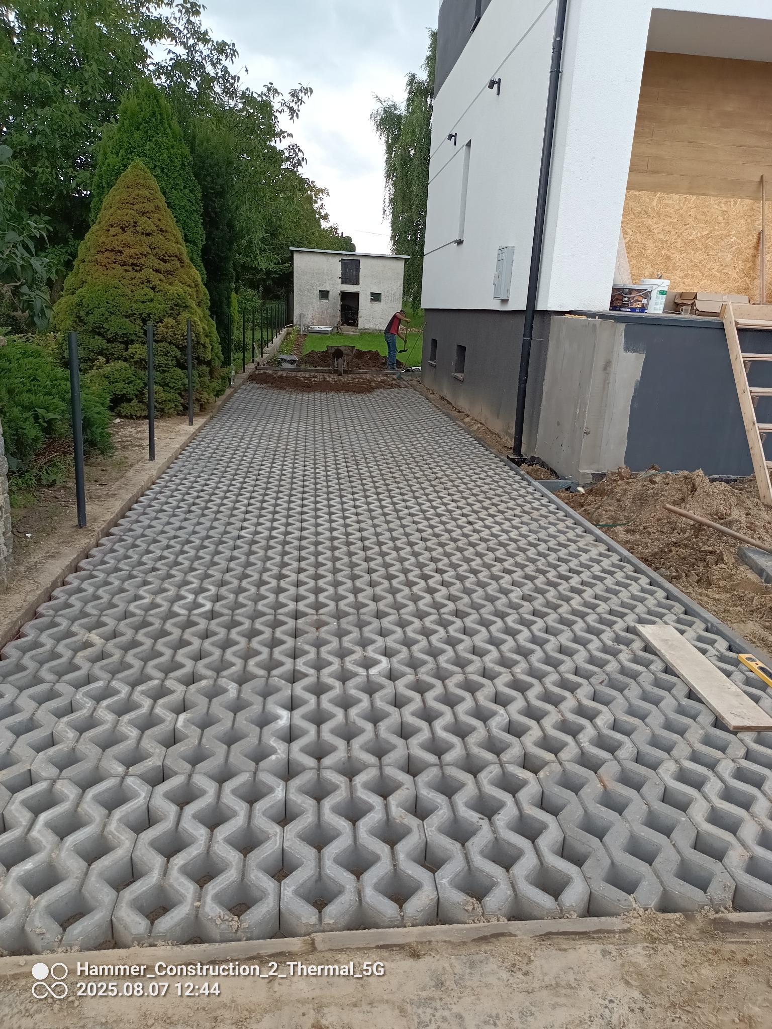Ułożona szara kostka ażurowa na podjeździe, widoczny fragment budynku i prace ziemne w tle. Kostka ułożona równo, tworząca wzór. Prace brukarskie w toku.