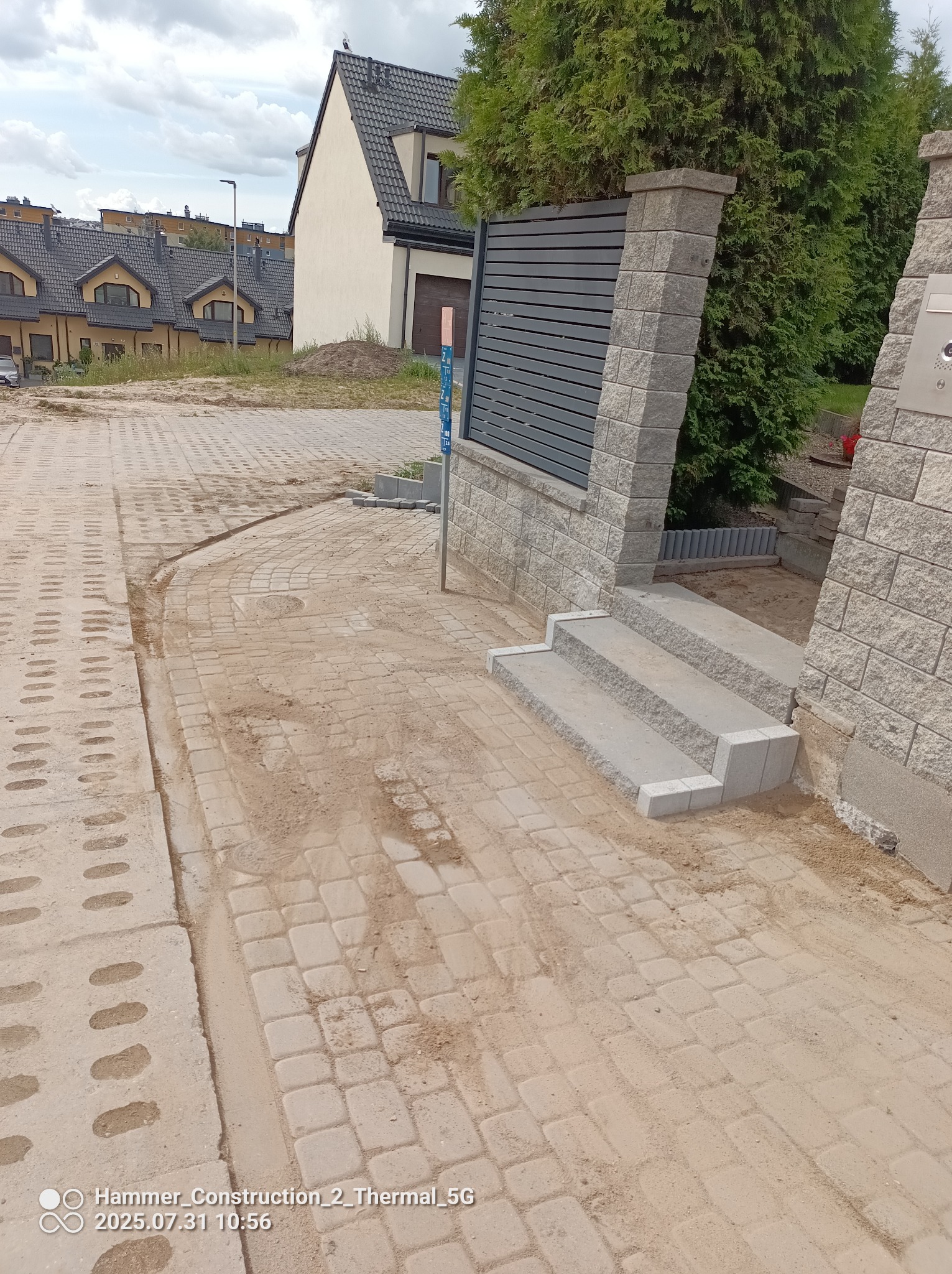 Ukończone podejście z kostki brukowej do posesji z trzema schodami i nowoczesnym ogrodzeniem z szarej cegły, na tle budynków mieszkalnych.