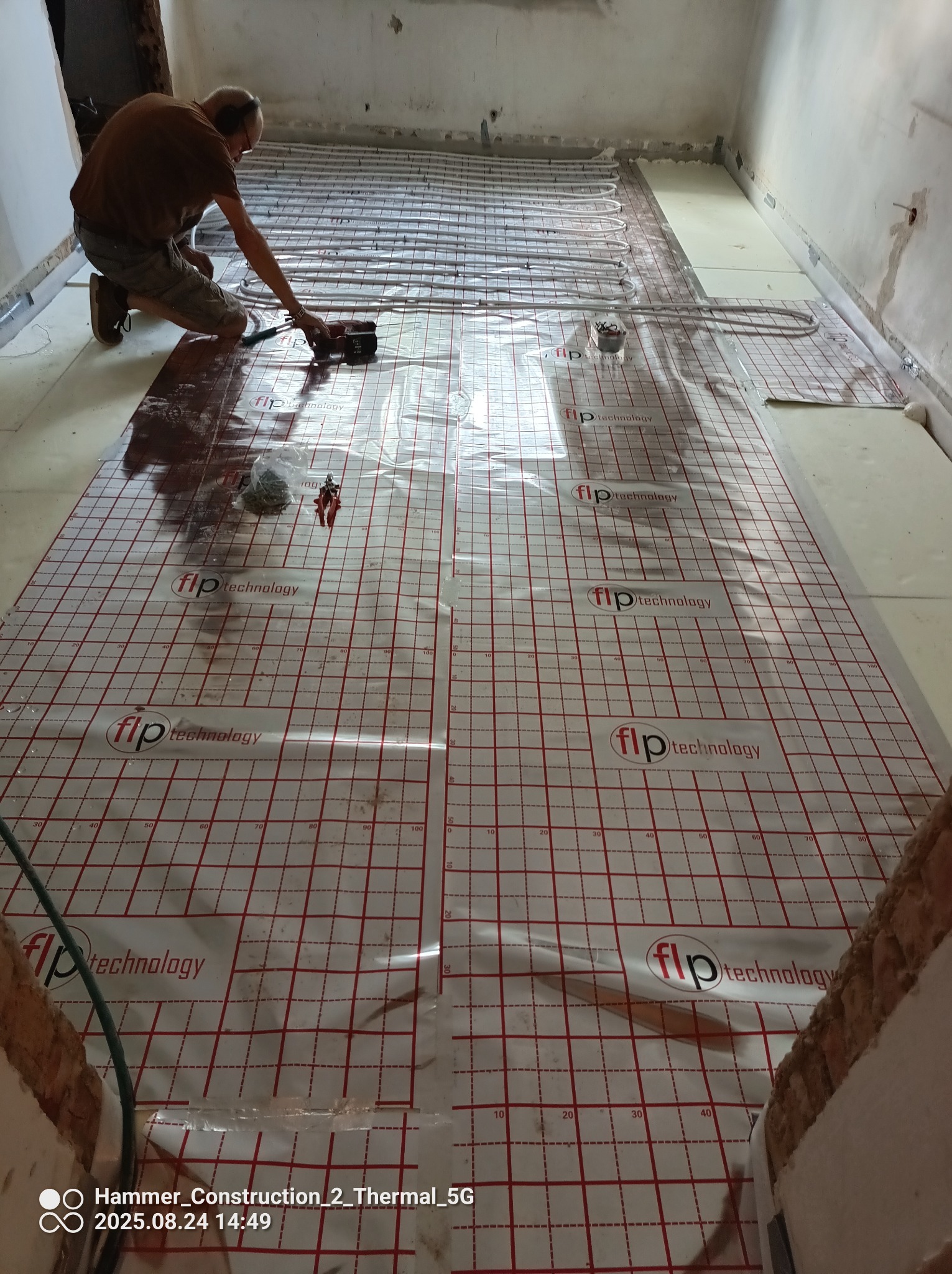 Montaż ogrzewania podłogowego: instalator mocuje rury do podłoża z folią rastrową. Praca w toku, widoczne narzędzia i elementy instalacji. Jasne wnętrze w trakcie remontu.