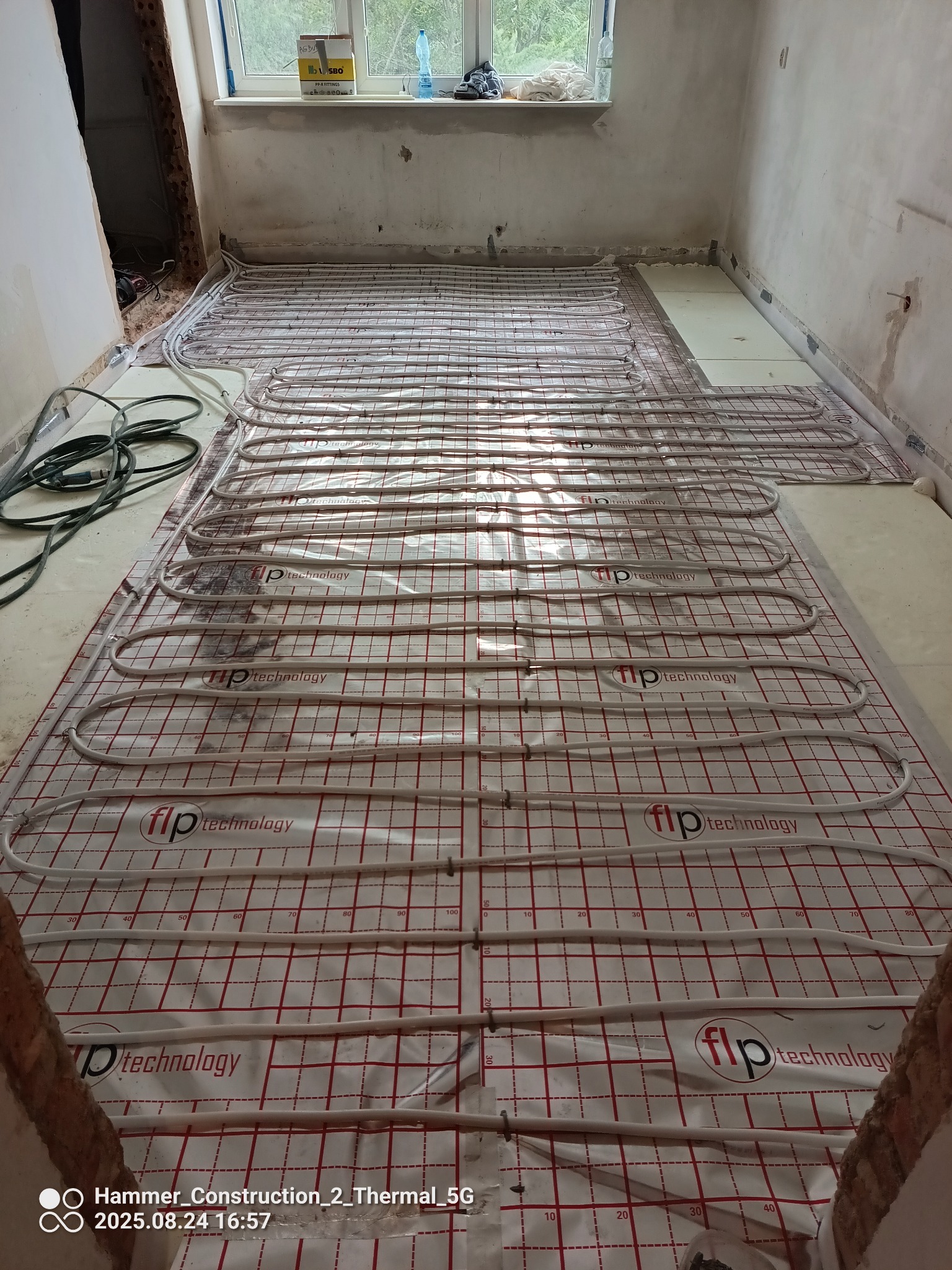 Instalacja ogrzewania podłogowego z białych rur na folii z czerwoną kratką w surowym wnętrzu remontowanego pomieszczenia. Widoczne kable i okno w tle.