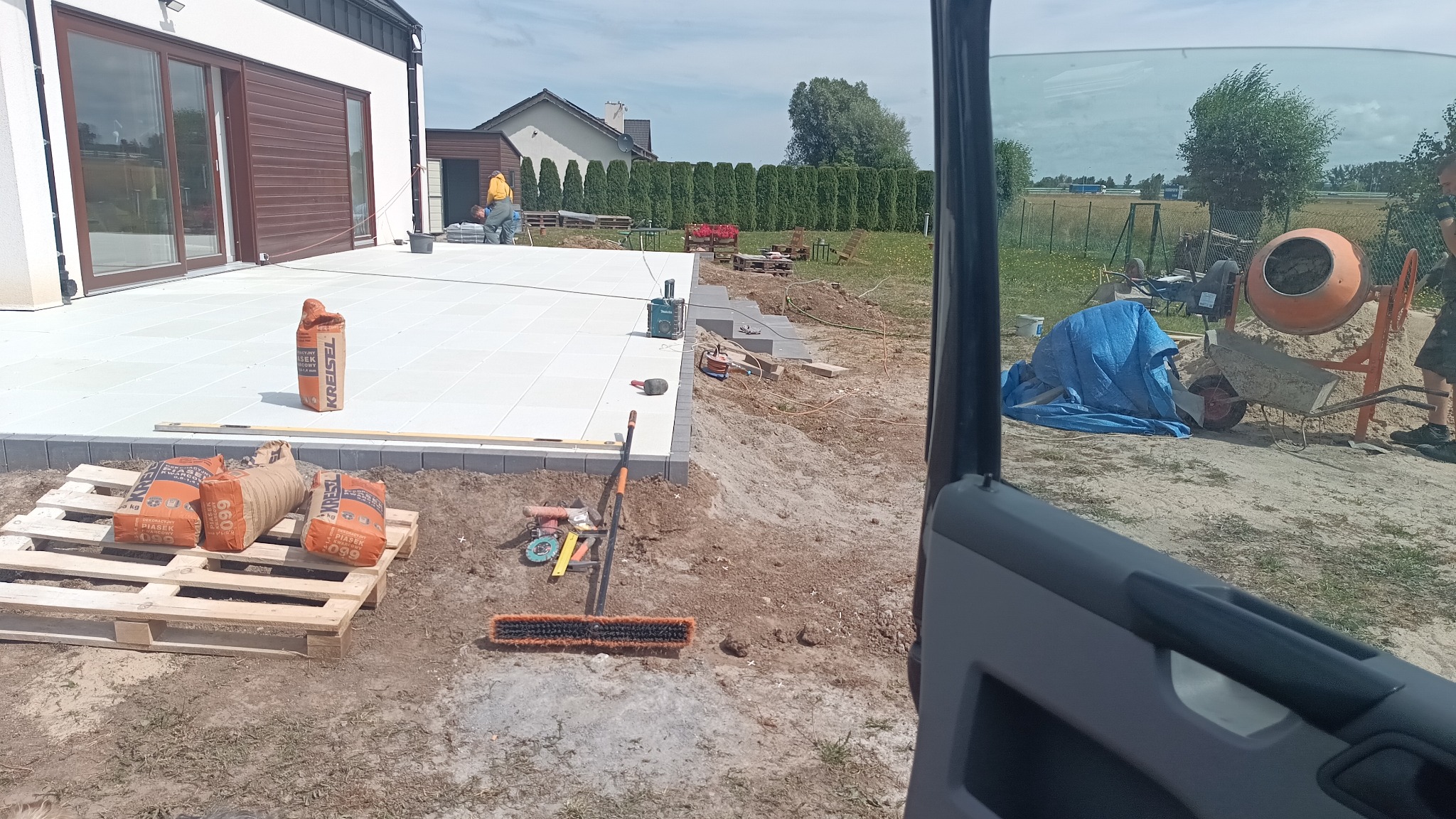 Jasnoszara kostka brukowa na tarasie w trakcie układania. Widoczne narzędzia, materiały budowlane i betoniarka. W tle dom z brązowymi elementami.