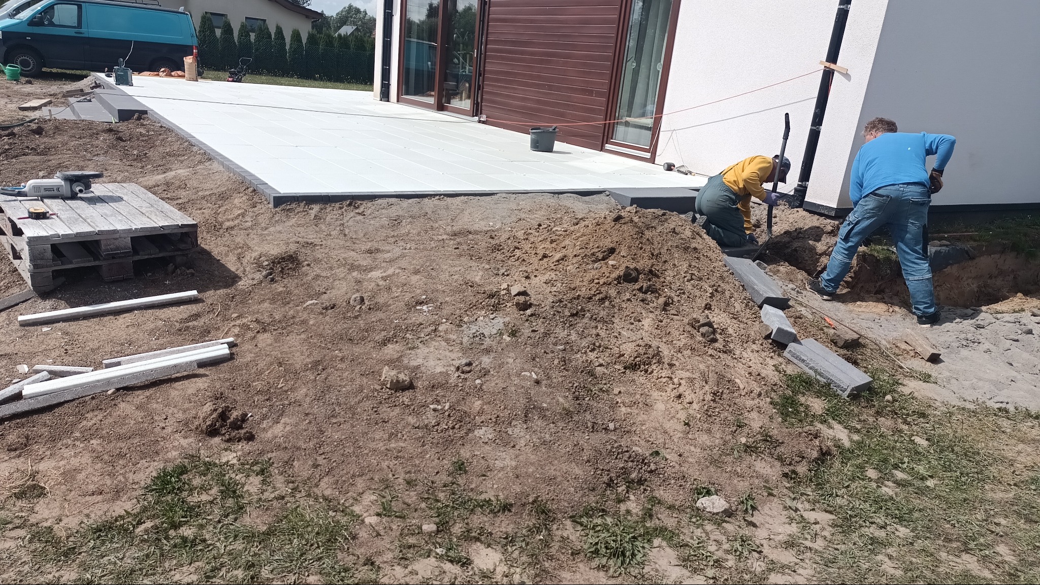 Montaż tarasu z jasnych płyt na gruncie. Dwóch pracowników układa obrzeża z szarej kostki. Widoczny paleta z narzędziami i materiały budowlane.