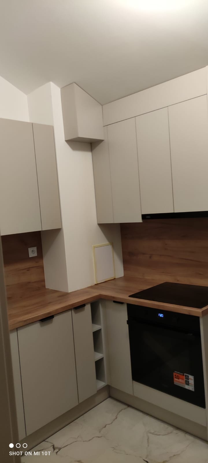 Jasna kuchnia z drewnianym blatem i zabudową. Widoczny piekarnik i szafki z szarymi frontami. Na ścianie gniazdko elektryczne i otwór rewizyjny.