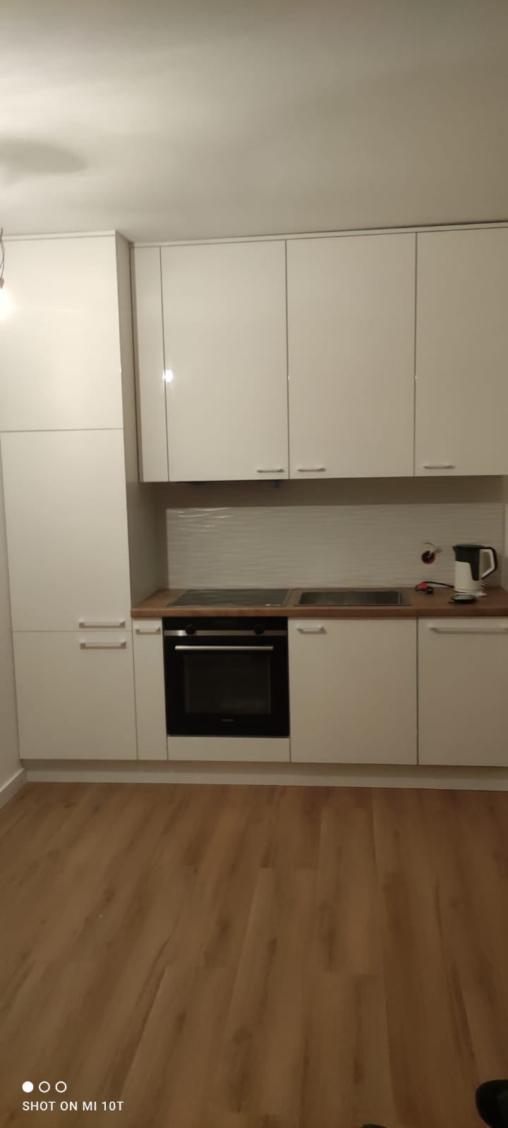 Biała, błyszcząca zabudowa kuchenna z drewnianym blatem, widok na płytę indukcyjną i piekarnik. Minimalistyczny styl, jasne wnętrze, drewniana podłoga.