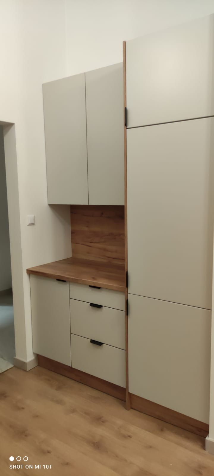 Nowoczesna zabudowa meblowa w odcieniach bieli i drewna, z minimalistycznymi uchwytami, tworząca spójną całość z drewnianą podłogą. Widok z perspektywy podłogi.