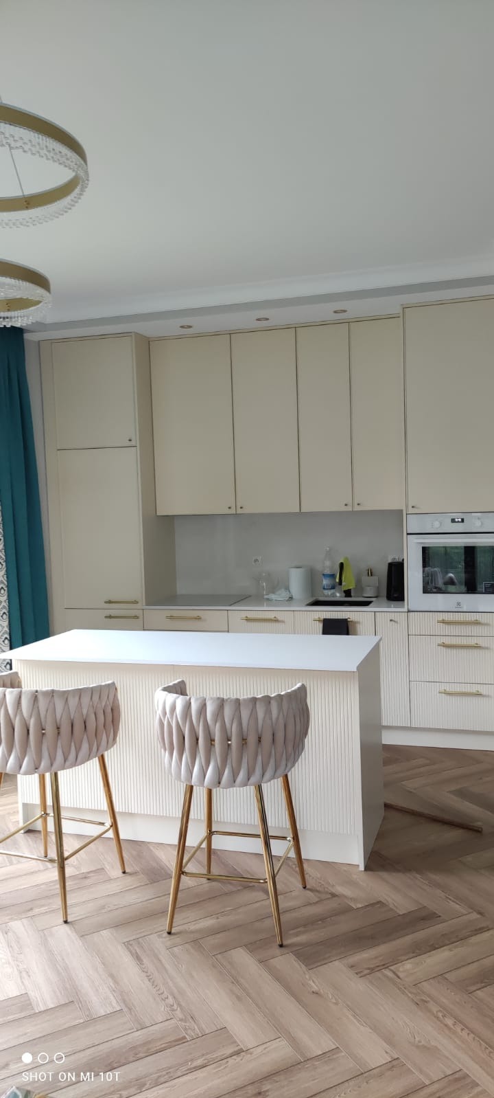 Beżowa kuchnia z wyspą, złote akcenty, krzesła barowe z plecionym oparciem, parkiet w jodełkę. Nowoczesne oświetlenie i minimalistyczny design.