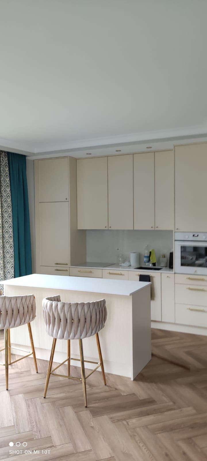Jasna kuchnia z wyspą i dwoma krzesłami barowymi. Drewniana podłoga w jodełkę. Kremowe szafki z delikatnymi złotymi uchwytami. Błękitne zasłony w tle.