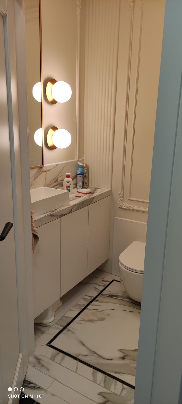 Elegancka łazienka w kremowych tonacjach: marmurowy blat, białe szafki pod umywalką, designerskie oświetlenie i marmurowe płytki podłogowe z czarną obwódką.