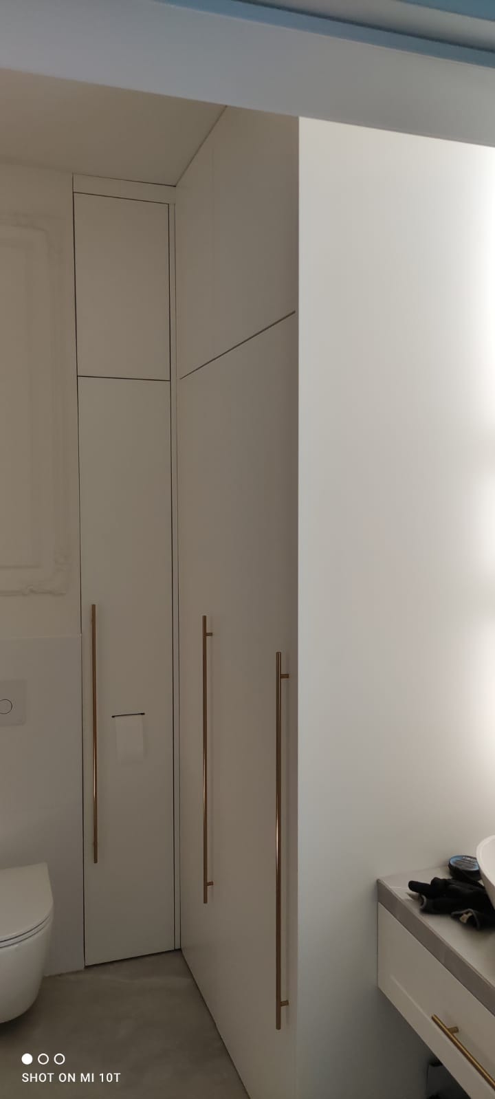 Biała zabudowa meblowa w łazience z długimi, złotymi uchwytami, widoczny fragment toalety i umywalki. Minimalistyczny design, jasne wnętrze.