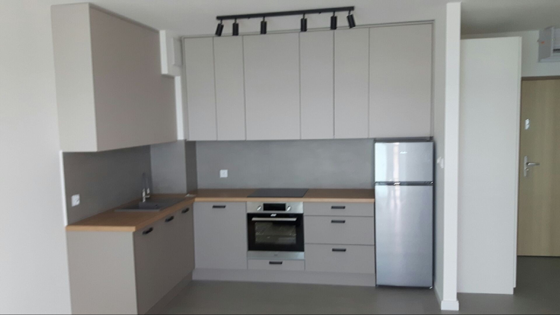 Jasnoszara kuchnia z drewnianym blatem, oświetleniem szynowym i srebrną lodówką. Widok na minimalistyczne wnętrze z frontami bez uchwytów.