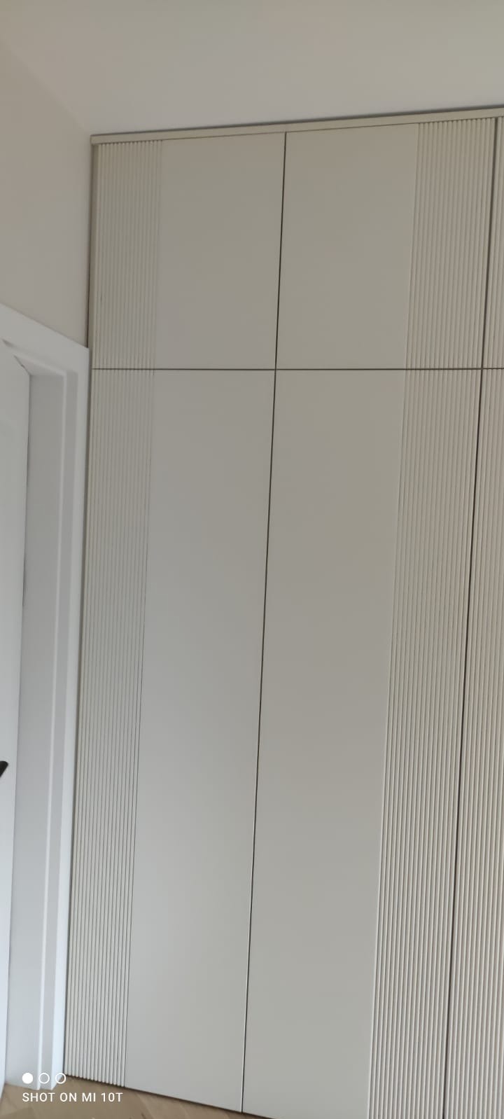 Biała szafa wnękowa z frezowanymi frontami, widoczna z bliska, prezentująca minimalistyczny design i precyzję wykonania. Element wystroju wnętrza.