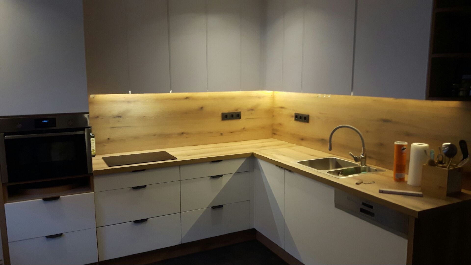 Nowoczesna kuchnia w kształcie litery L z białymi szafkami i drewnianym blatem, oświetlona od góry, z widocznym zlewem i piekarnikiem.