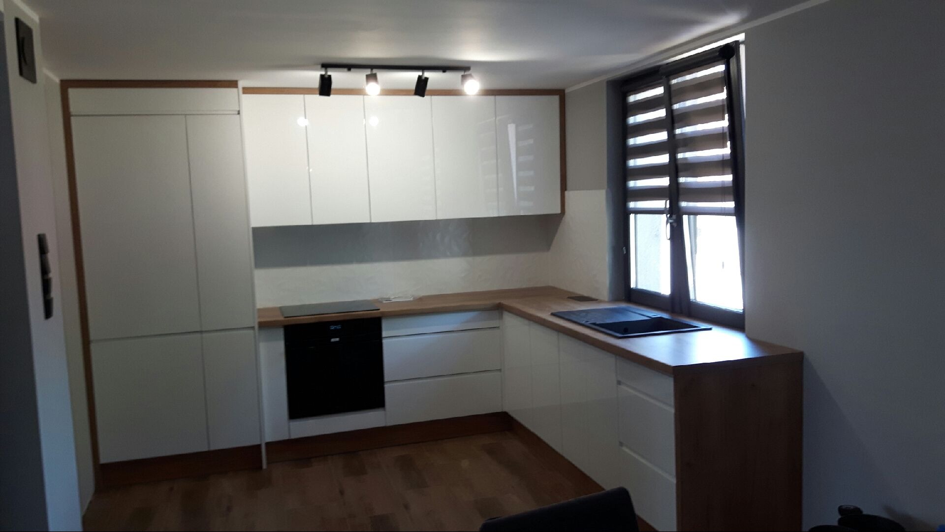 Nowoczesna kuchnia z białymi, lakierowanymi frontami i drewnianymi blatami. Minimalistyczne oświetlenie punktowe i okno z żaluzjami. Widok na zabudowę kuchenną.