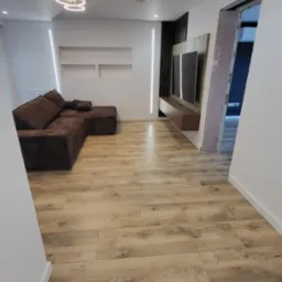 Nowoczesny salon z parkietem w odcieniach drewna, brązową sofą narożną i podświetlaną ścianą TV. Widoczne oświetlenie sufitowe i fragment otwartego przejścia.