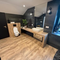RAS-BUD Dawid Rasiński - Nowoczesna łazienka z drewnianą podłogą i blatem, grafitowe płytki ścienne, owalna umywalka, duże lustro i toaleta. Minimalistyczny design, roślina w oknie.