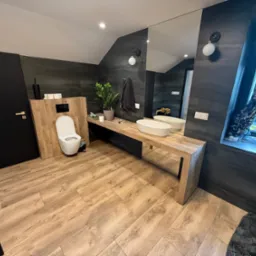 Nowoczesna łazienka z drewnianą podłogą i blatem, grafitowe płytki ścienne, owalna umywalka, duże lustro i toaleta. Minimalistyczny design, roślina w oknie.