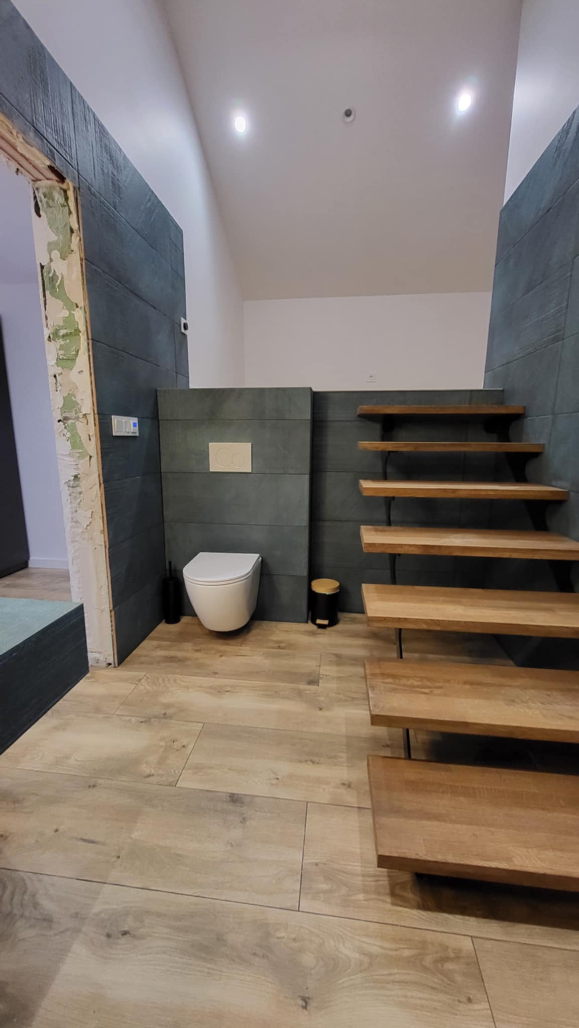 Nowoczesna łazienka z drewnianymi schodami, szarą glazurą i podwieszaną toaletą. Częściowo wykończone obramowanie drzwi.