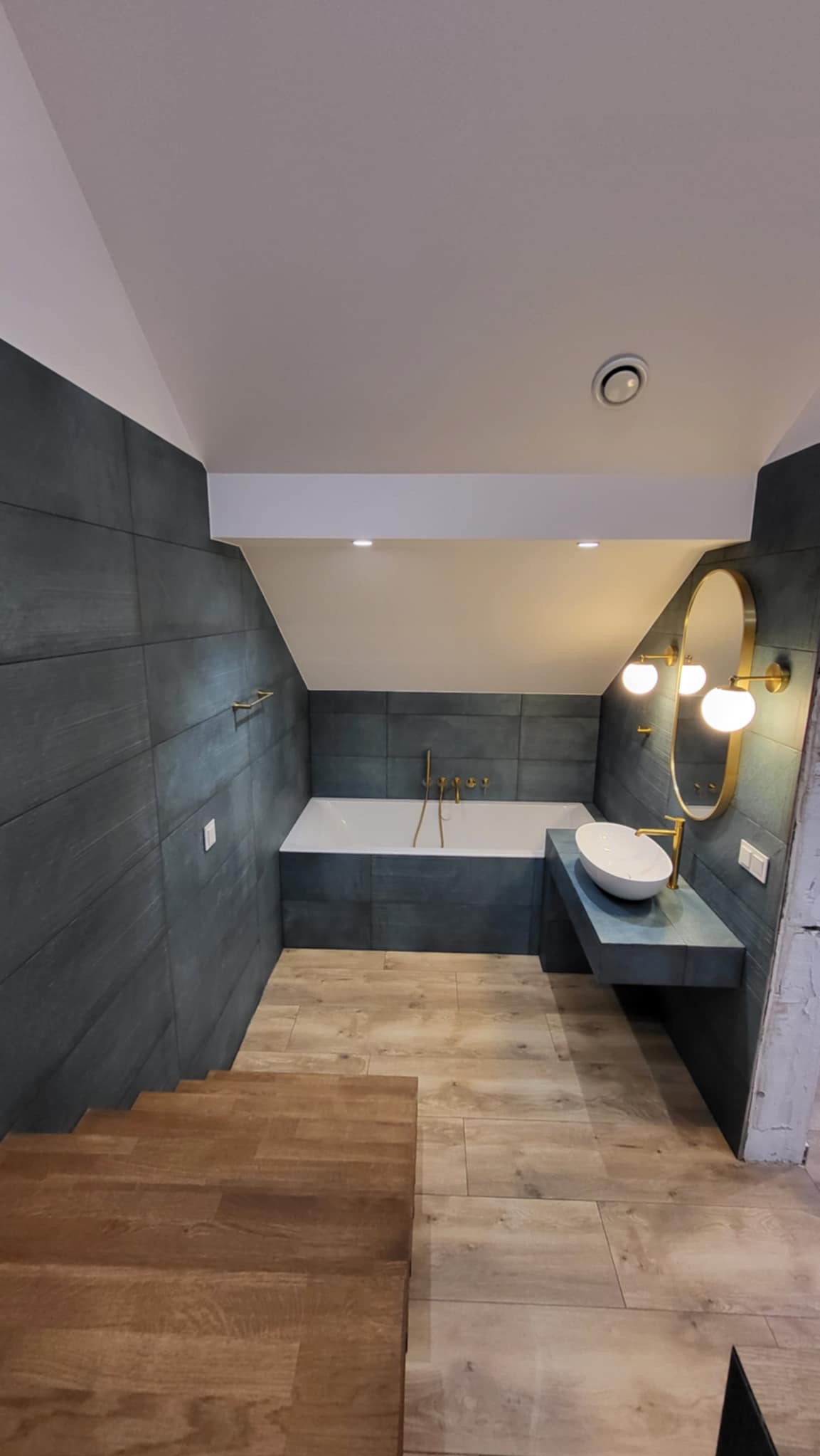 Nowoczesna łazienka na poddaszu: grafitowe płytki, drewnopodobna podłoga, biała wanna i umywalka z złotymi detalami. Oświetlenie punktowe i owalne lustro.
