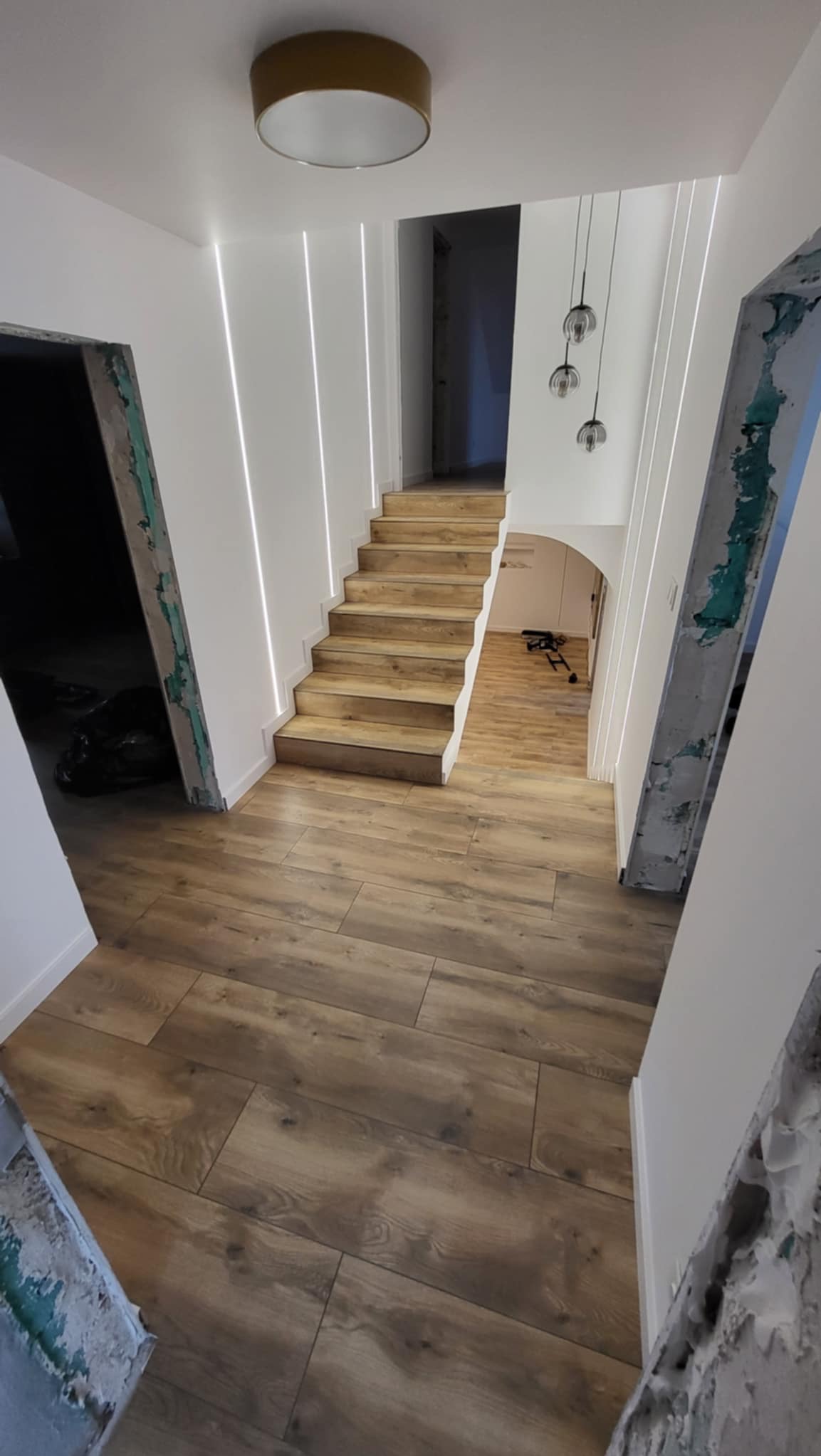 Wykończone schody z drewnianymi stopniami i podświetleniem LED na ścianie, drewnopodobna podłoga, widoczne ślady prac wykończeniowych przy futrynach.