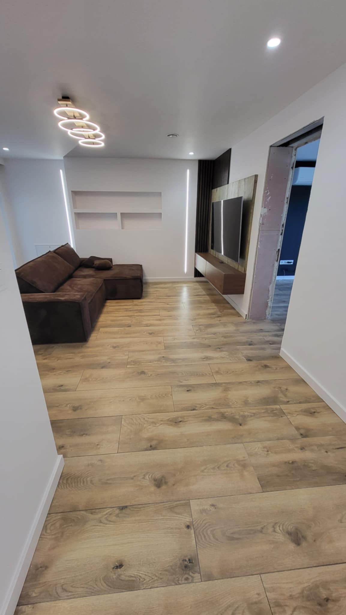 Nowoczesny salon z parkietem w odcieniach drewna, brązową sofą narożną i podświetlaną ścianą TV. Widoczne oświetlenie sufitowe i fragment otwartego przejścia.