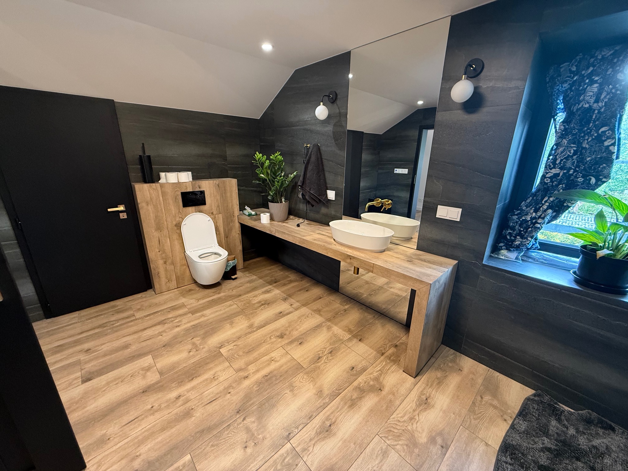 Nowoczesna łazienka z drewnianą podłogą i blatem, grafitowe płytki ścienne, owalna umywalka, duże lustro i toaleta. Minimalistyczny design, roślina w oknie.