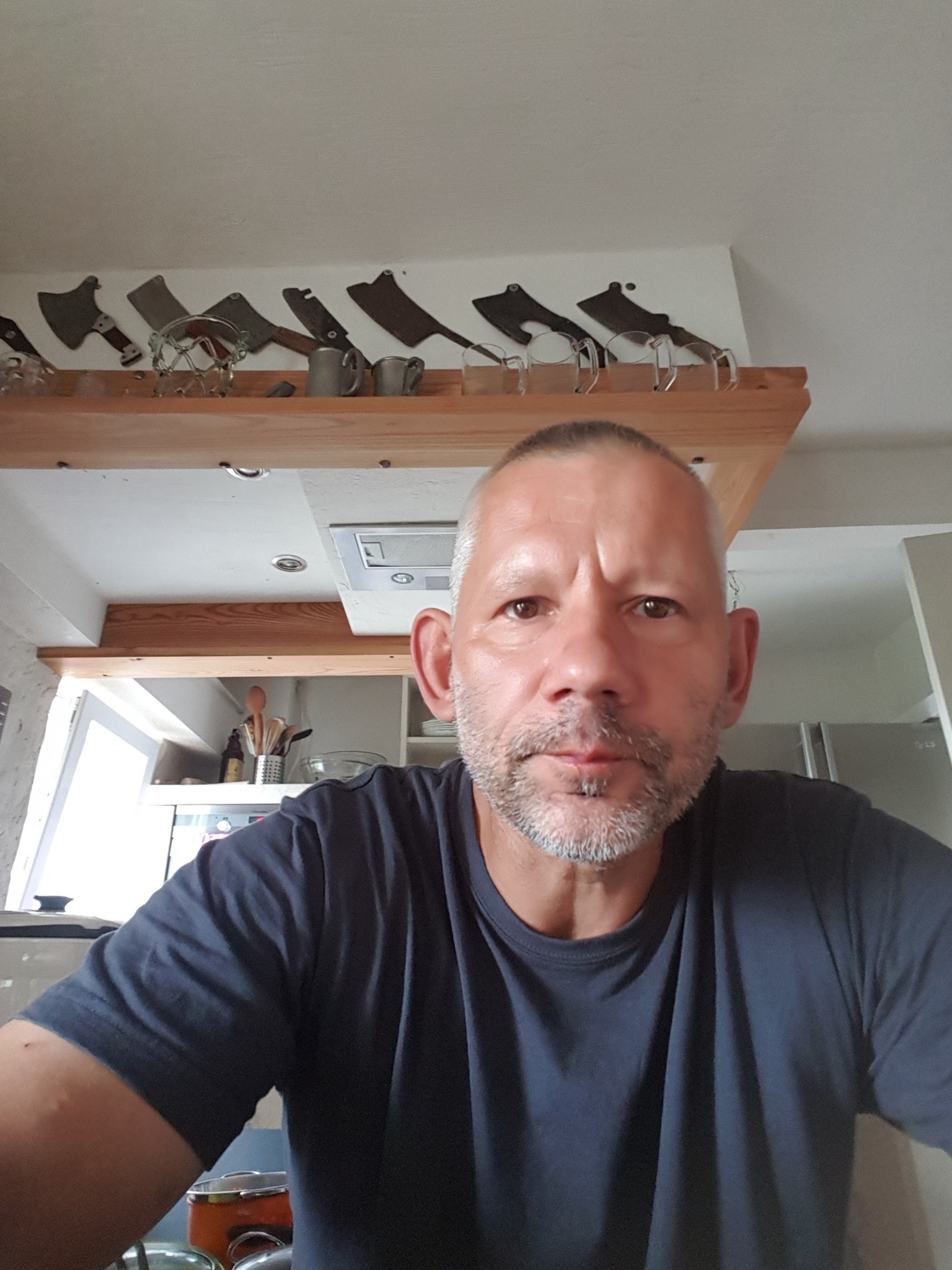 Mężczyzna w kuchni z kolekcją tasaków na półce, oświetlenie sufitowe i okno w tle. Ujęcie z dołu.