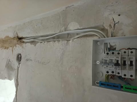 Otwarta skrzynka elektryczna z zabezpieczeniami na surowej ścianie, widoczne kable w bruździe, etap prac wykończeniowych.