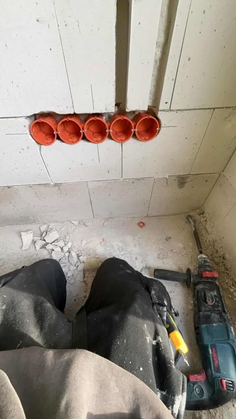 Rząd pomarańczowych puszek elektrycznych w ścianie z betonu komórkowego, obok leży wiertarka udarowa i fragment ubrania osoby wykonującej instalację.