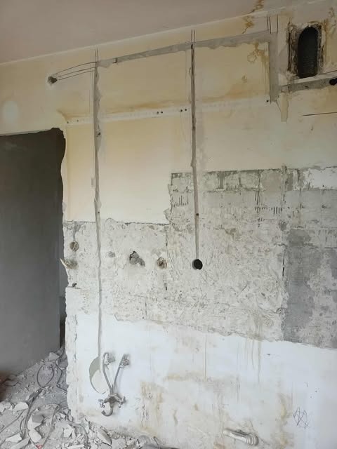Ściana w trakcie remontu z widocznymi przewodami elektrycznymi i instalacją wodną, gruz na podłodze. Widoczne ślady po kuciu w ścianie.