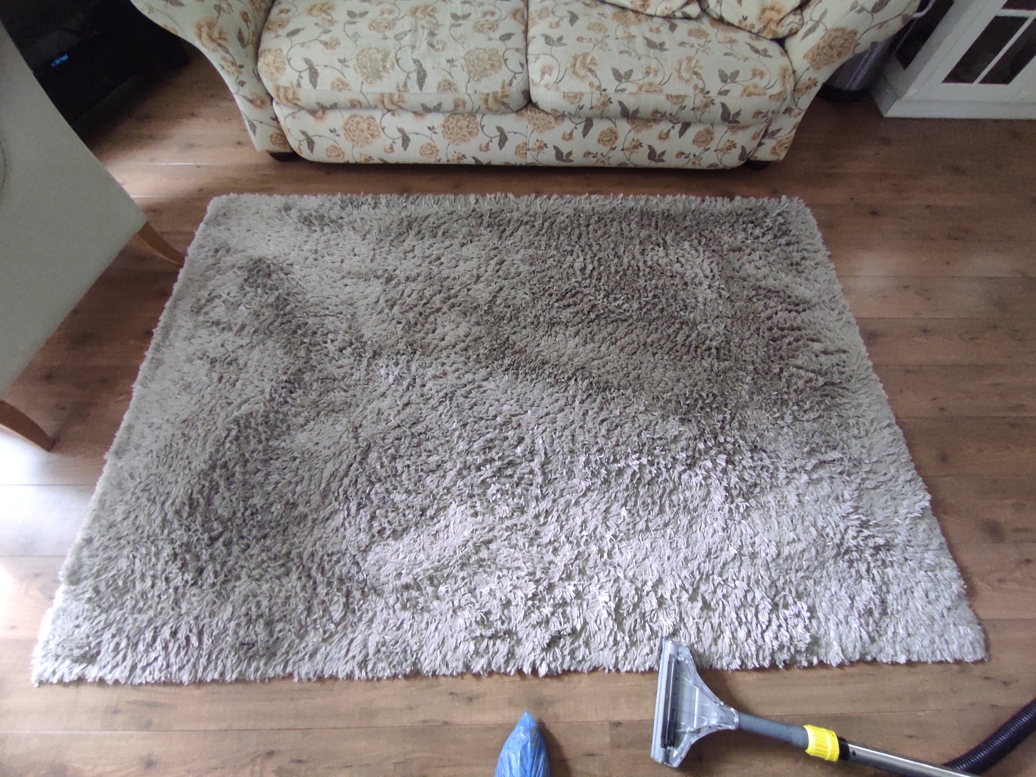 Jasnoszary dywan shaggy leżący na drewnianej podłodze, obok sofy w kwiaty i dyszy odkurzacza piorącego z niebieską osłoną. Widok z góry.
