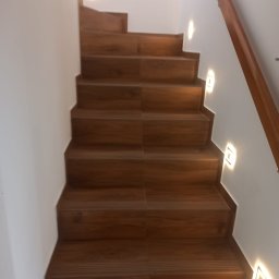 Usługi Wykończeniowe - Drewniane schody z oświetleniem punktowym na ścianie, widok z dołu. Poręcz z drewna po prawej stronie. Minimalistyczny design.