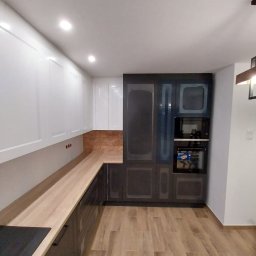 Usługi Wykończeniowe - Nowoczesna kuchnia z białymi szafkami górnymi i ciemnymi dolnymi, drewnianym blatem oraz podłogą imitującą drewno. W zabudowie piekarnik i mikrofalówka.