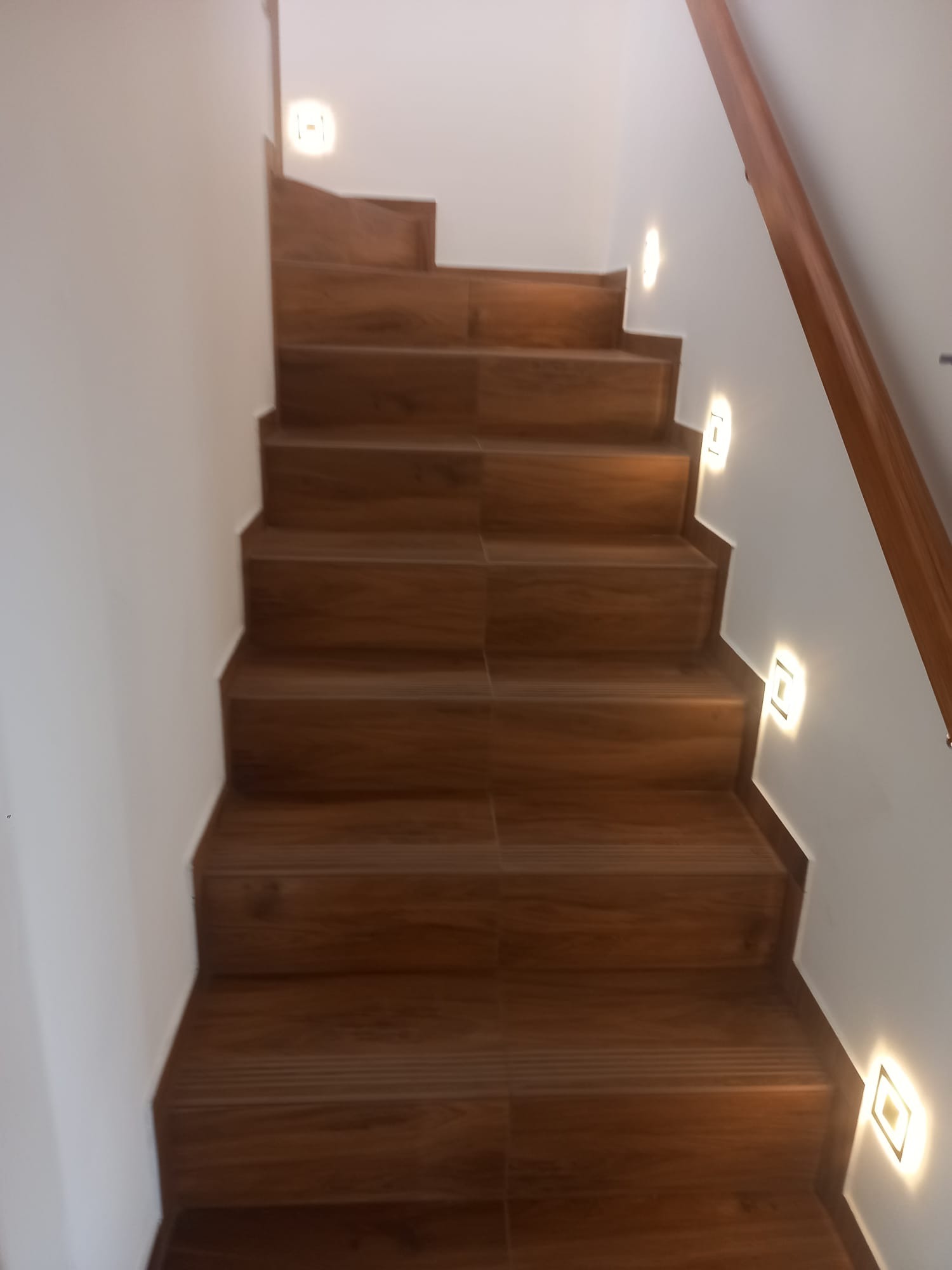 Drewniane schody z oświetleniem punktowym na ścianie, widok z dołu. Poręcz z drewna po prawej stronie. Minimalistyczny design.