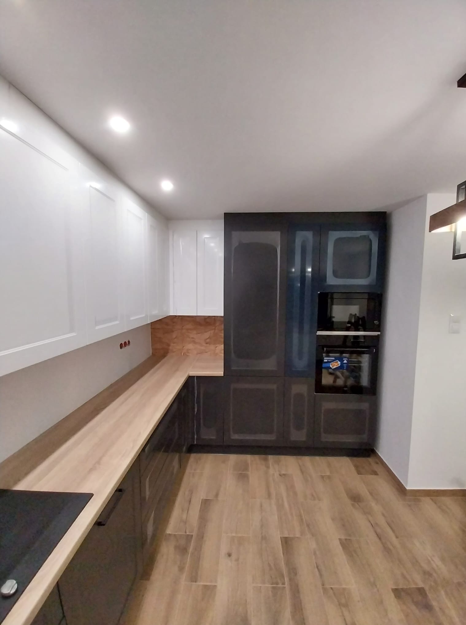 Nowoczesna kuchnia z białymi szafkami górnymi i ciemnymi dolnymi, drewnianym blatem oraz podłogą imitującą drewno. W zabudowie piekarnik i mikrofalówka.