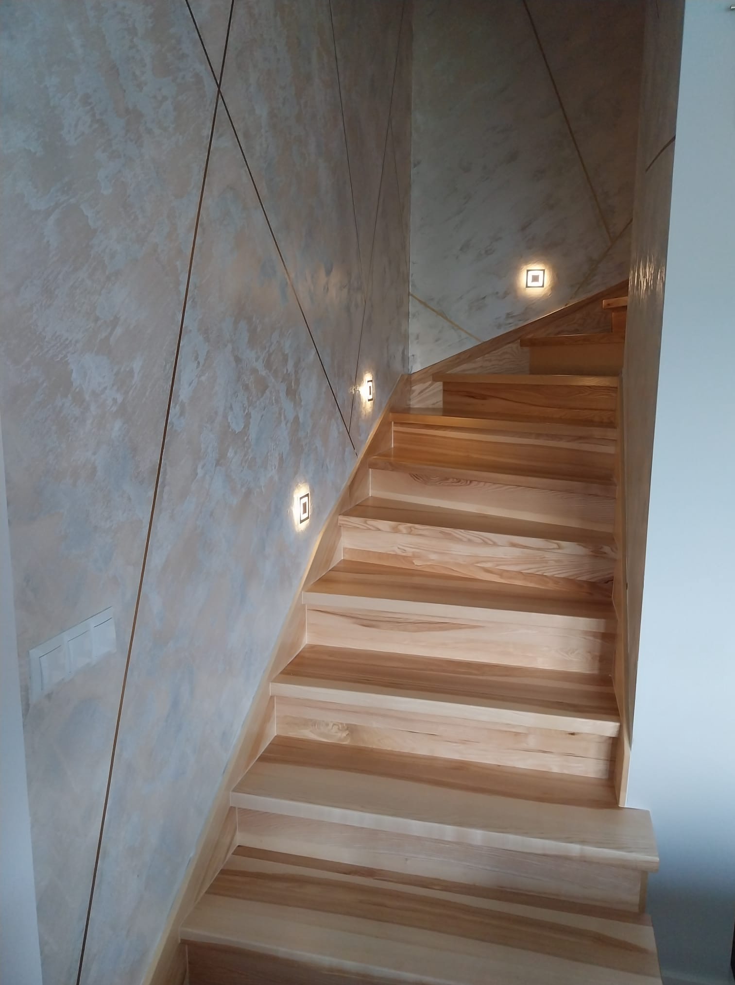 Drewniane schody z oświetleniem punktowym na ścianie o dekoracyjnej fakturze i geometrycznych liniach. Widoczne włączniki światła.
