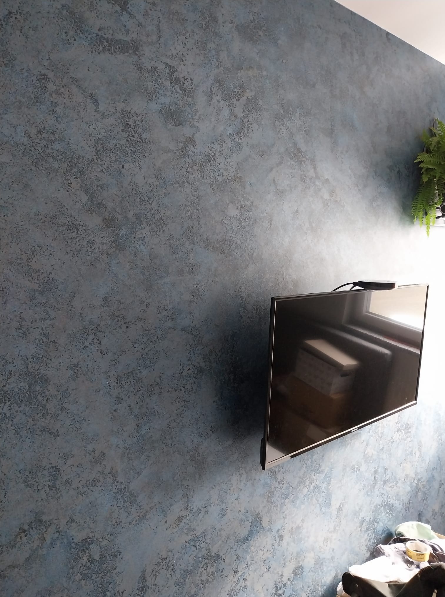 Ściana z dekoracyjnym, niebiesko-szarym tynkiem strukturalnym, obok wiszący telewizor i roślina doniczkowa. Widoczne niedoskonałości i nierówności faktury.
