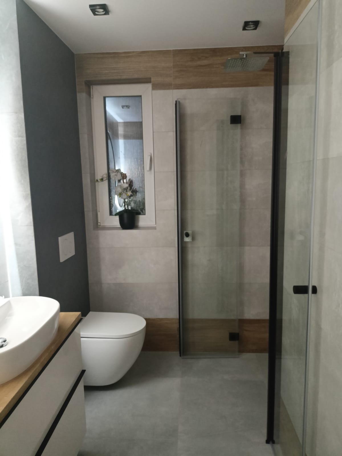 Nowoczesna łazienka z kabiną prysznicową typu walk-in, umywalką nablatową i toaletą. Ciekawa kompozycja płytek w odcieniach szarości i drewna. Minimalistyczny design.