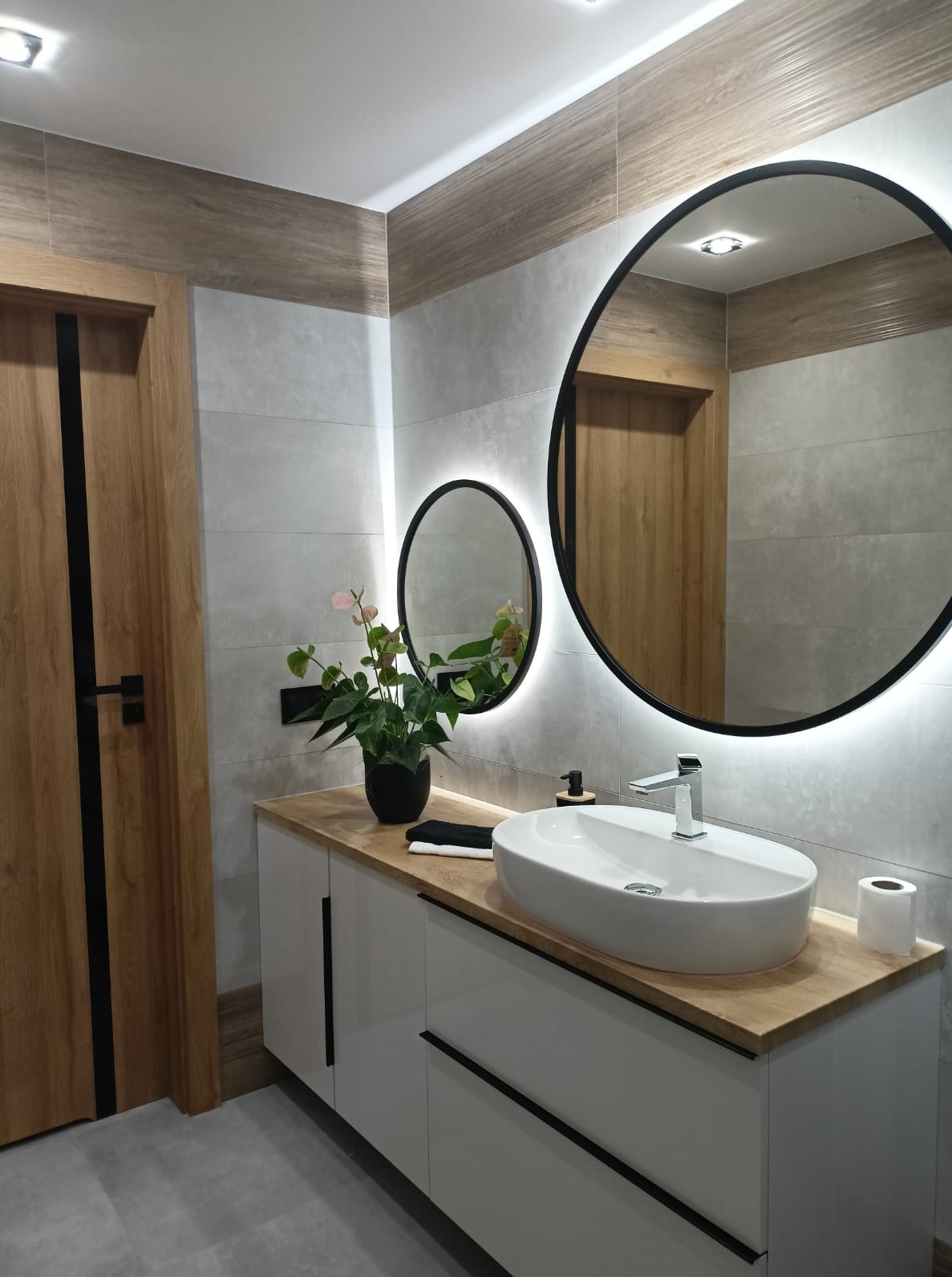 Nowoczesna łazienka z owalną umywalką nablatową, drewnianym blatem, podświetlanymi lustrami i drzwiami z drewnianym fornirem i czarnymi akcentami.