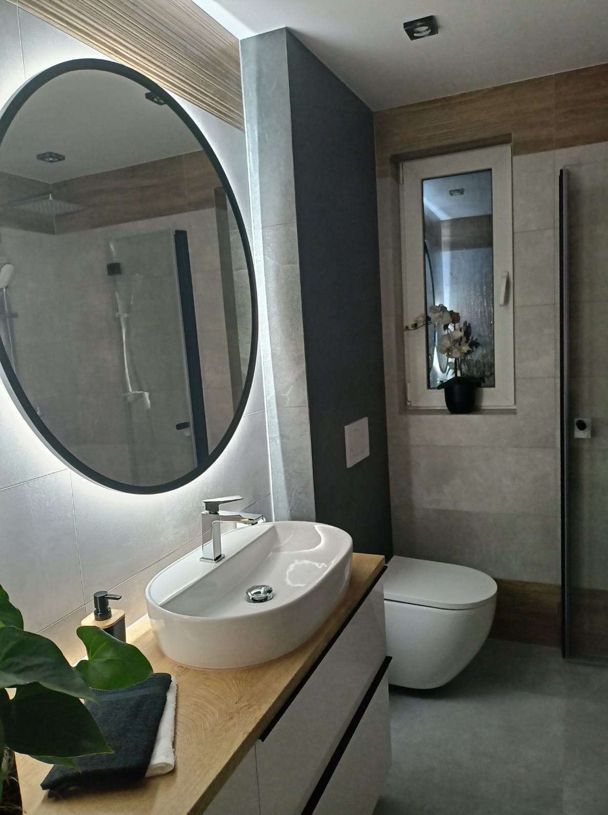 Nowoczesna łazienka po remoncie: owalne lustro z podświetleniem LED, umywalka nablatowa na drewnianym blacie, szare płytki ścienne, okno z kwiatami.