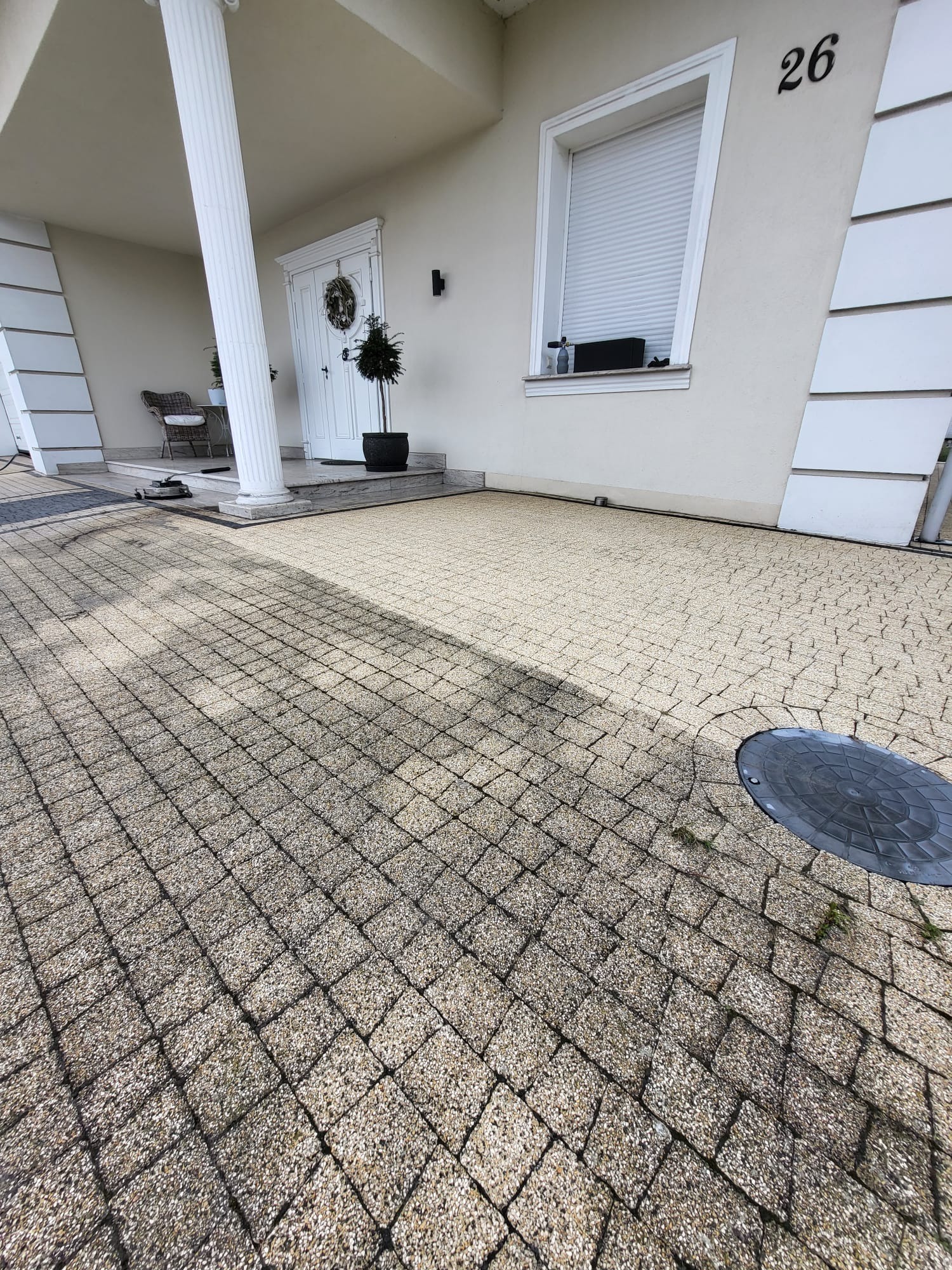 Jasna kostka brukowa przed domem z kolumnami, widoczny fragment czyszczonej powierzchni i studzienka kanalizacyjna. Numer domu 26.