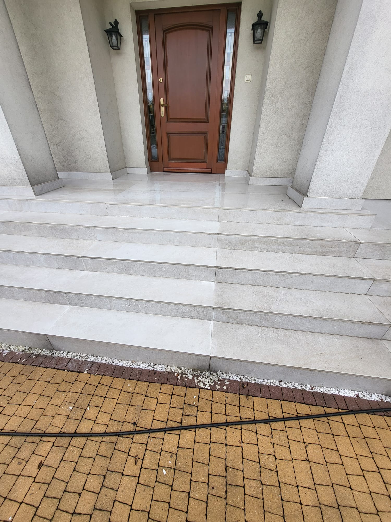 Wyślizgana, jasna kostka schodowa prowadząca do brązowych drzwi wejściowych, z widoczną kostką brukową w odcieniach brązu i czarnym wężem ogrodowym w dolnej części kadru.