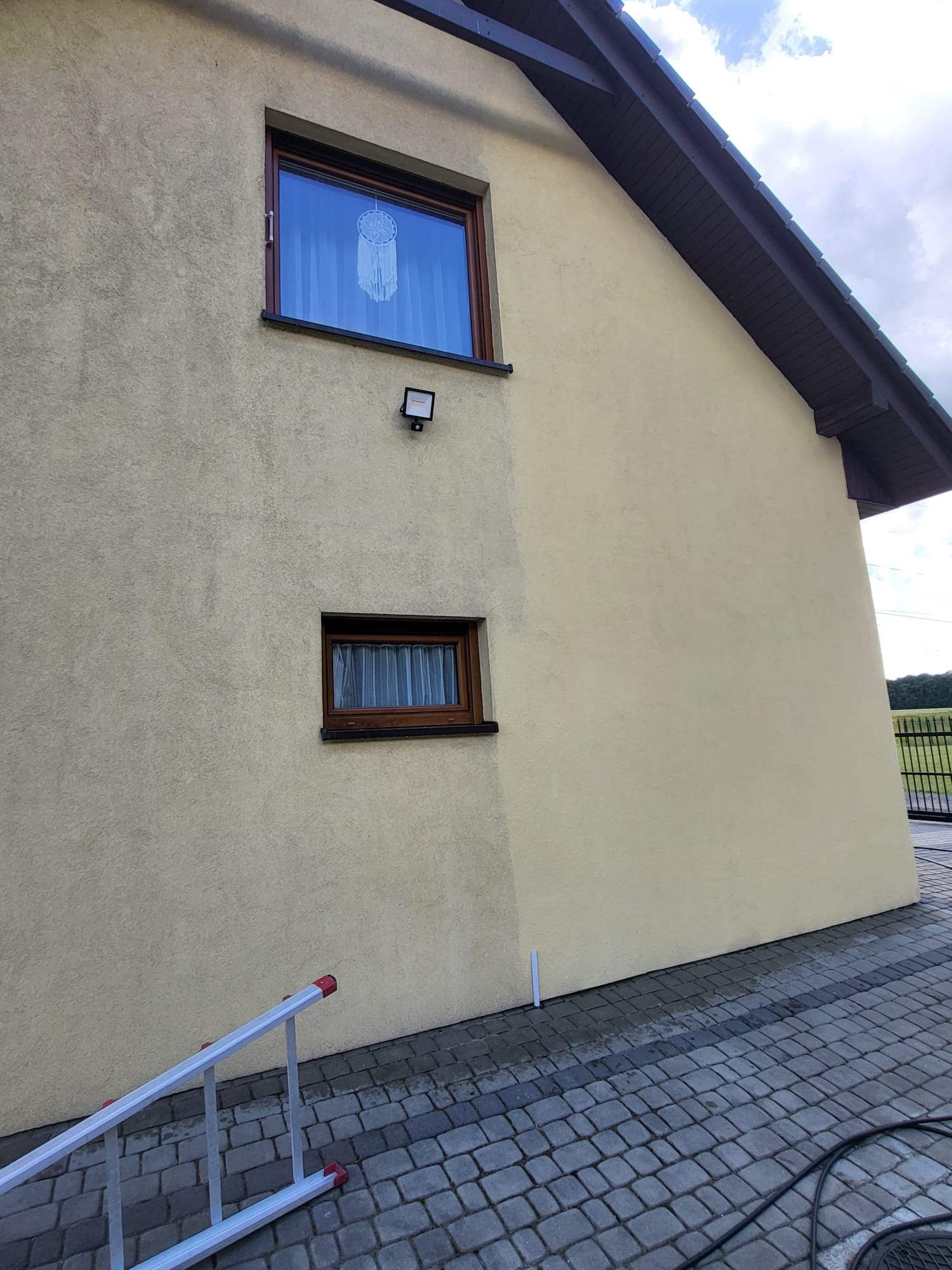 Fragment elewacji budynku po myciu ciśnieniowym, widoczna różnica w czystości. Częściowo widoczna drabina i kostka brukowa.