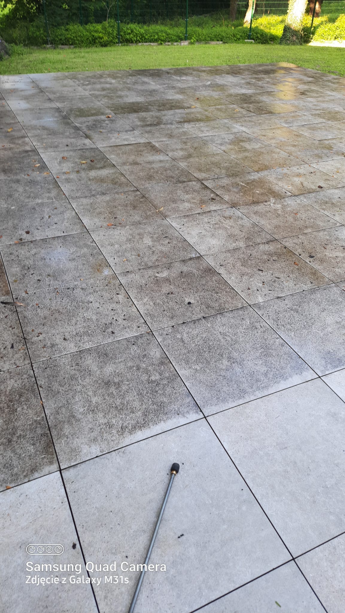 Fragment wyczyszczonej kostki brukowej w Legnicy, widoczna różnica między umytą a zabrudzoną powierzchnią. W tle trawnik i drzewa, dysza urządzenia do czyszczenia.