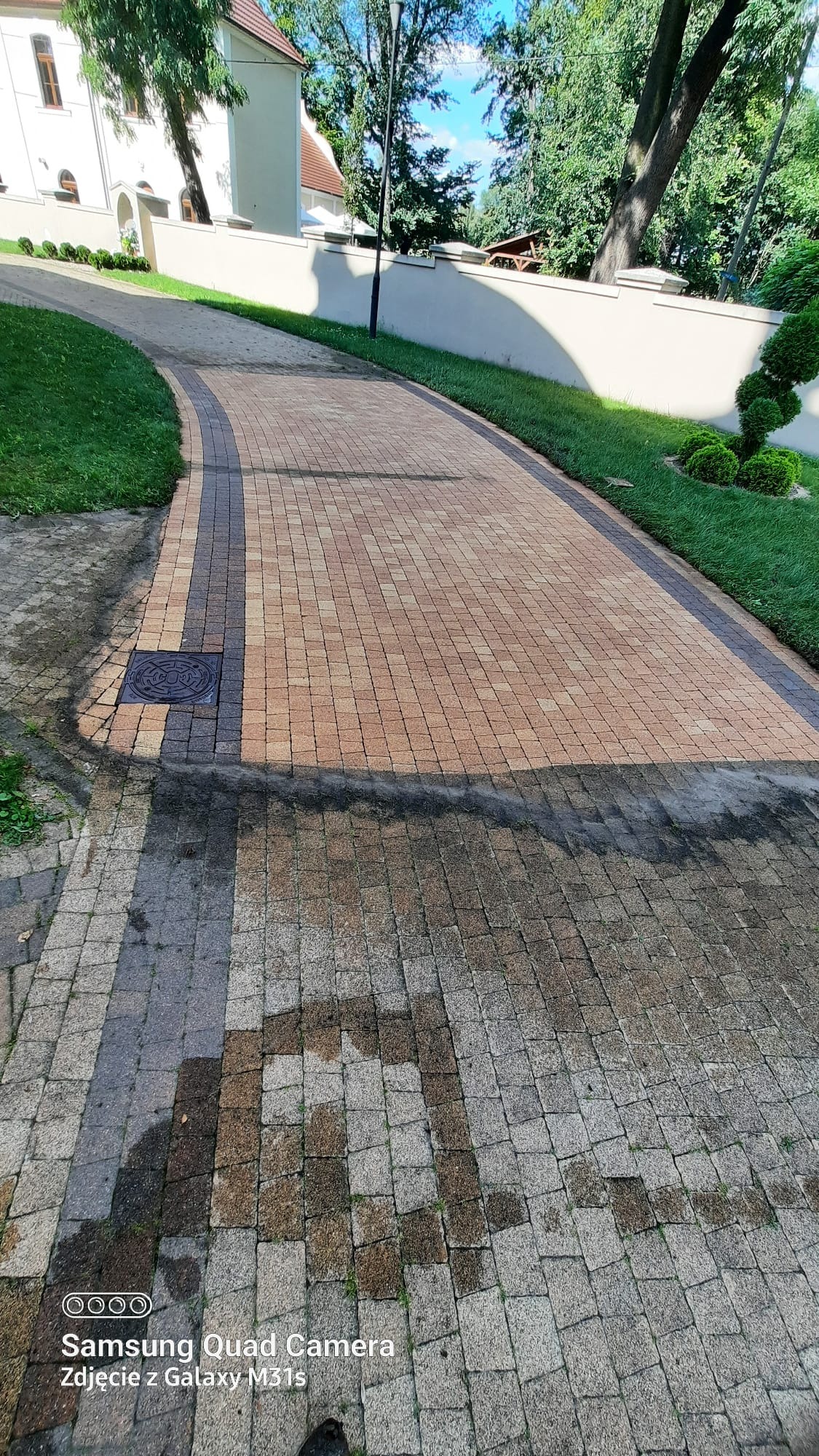 Częściowo umyta kostka brukowa z widocznym efektem czyszczenia, zarys czystej powierzchni na tle zabrudzonej. Widok na posesję z zielenią.