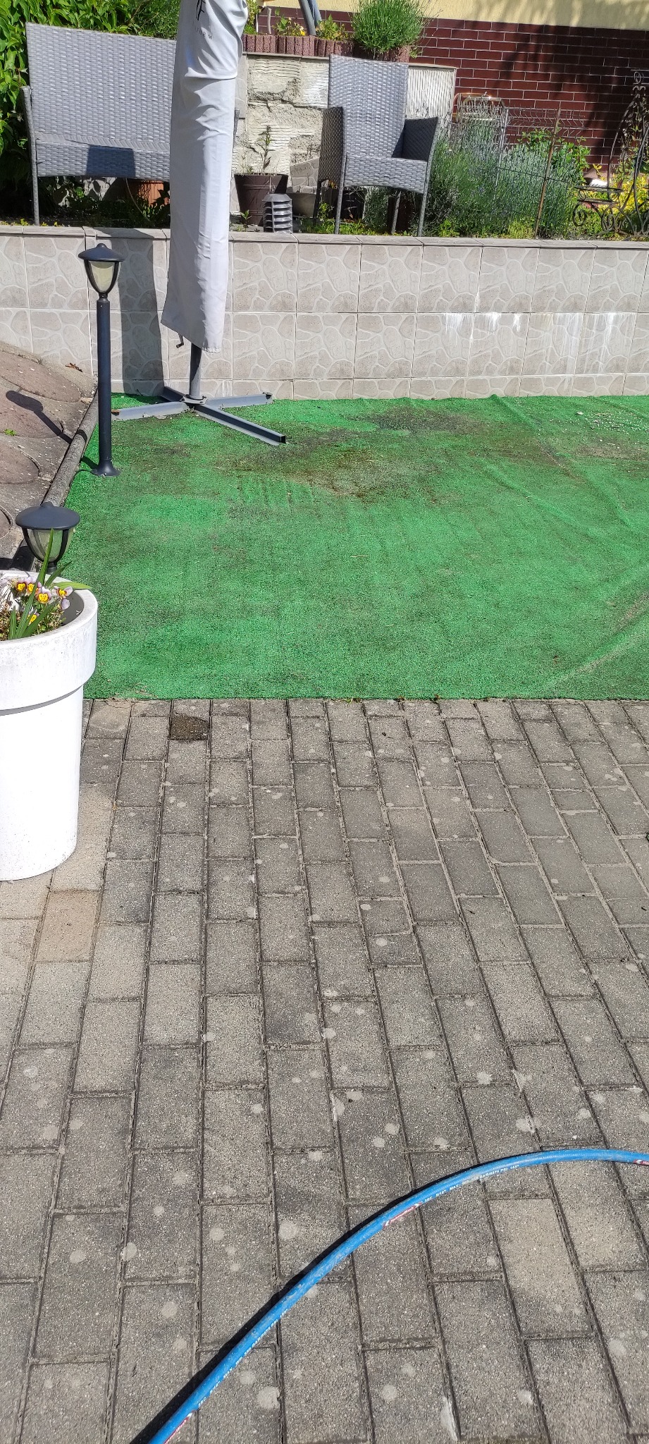 Brudna kostka brukowa i sztuczna trawa przed czyszczeniem. Widoczny wąż ogrodowy, donica z kwiatami, lampa ogrodowa i meble ogrodowe.