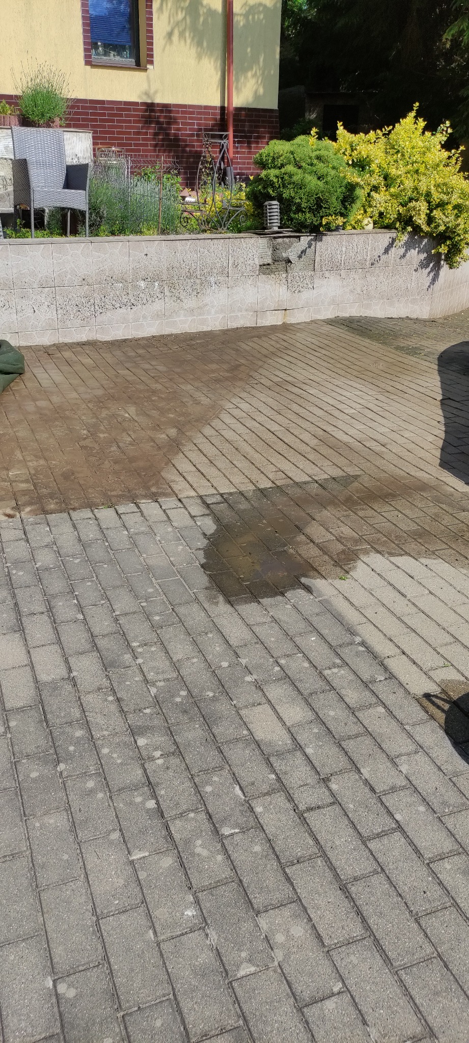 Częściowo umyta kostka brukowa na podjeździe, widoczna różnica w kolorze, tło stanowi budynek z ogrodem. Ujęcie z góry, w słoneczny dzień.