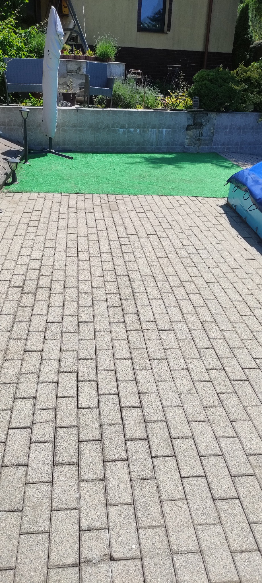 Jasnoszara kostka brukowa na posesji z zielonym trawnikiem i elementami architektury ogrodowej w tle, w słoneczny dzień.