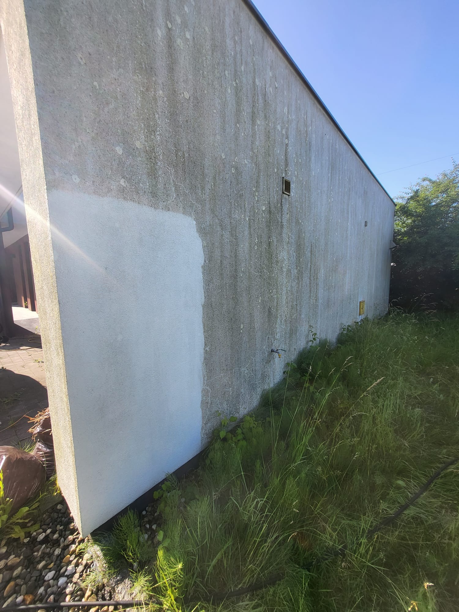 Fragment elewacji budynku przed i po czyszczeniu, widoczne kontrasty zabrudzeń i czystej, jasnej powierzchni. Wysoka trawa i kamienie wzdłuż ściany.
