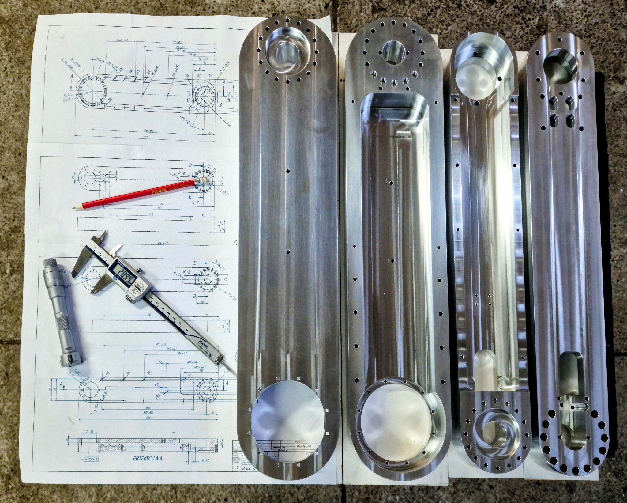 Precyzyjnie obrobione metalowe elementy na rysunku technicznym z ołówkiem i narzędziami pomiarowymi. Widoczne otwory i frezowania.