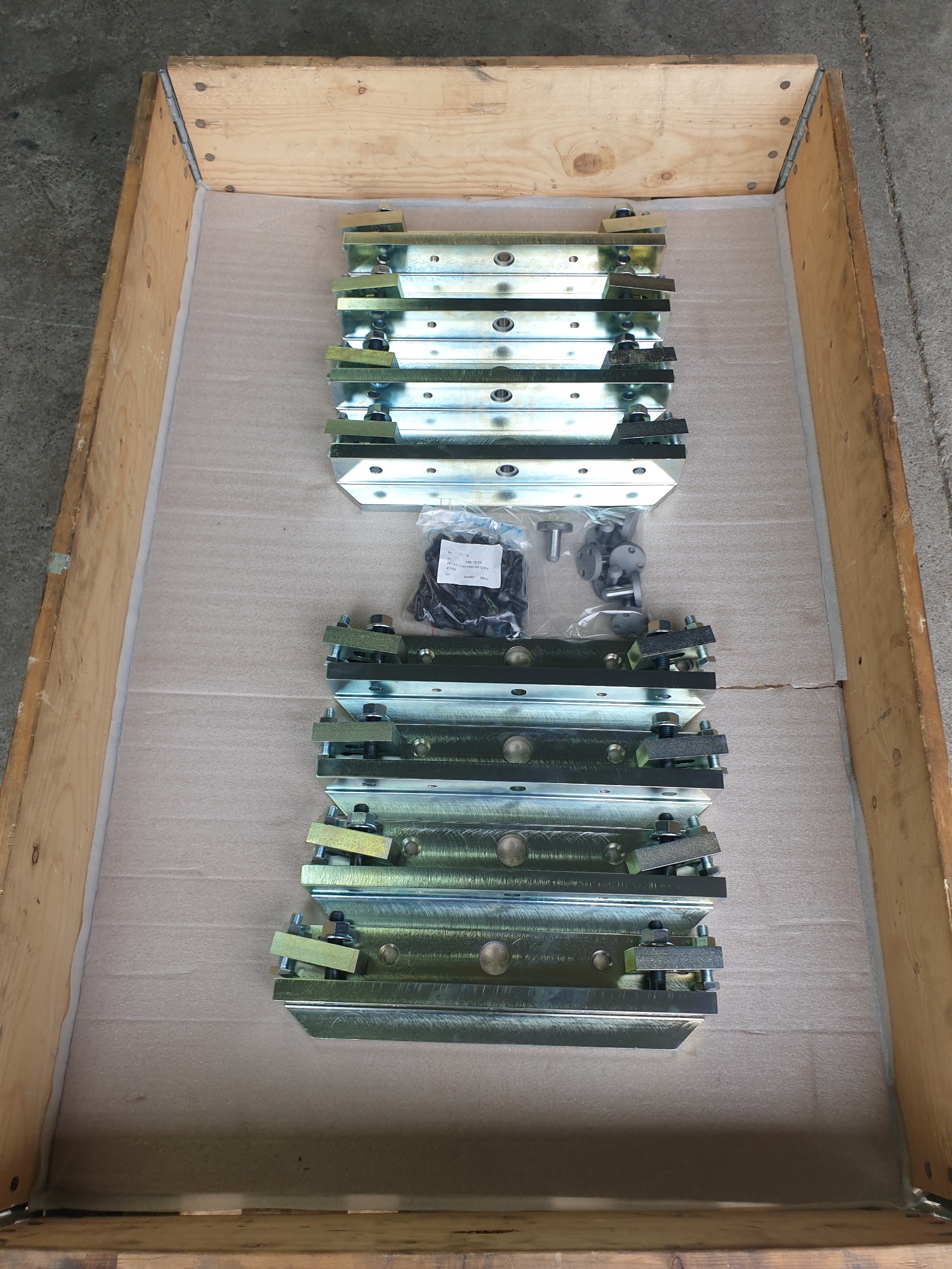 Metalowe elementy z nakrętkami i śrubami w drewnianej skrzyni, gotowe do montażu. Widoczne detale obróbki i wykończenia. Części ułożone równo na kartonie.